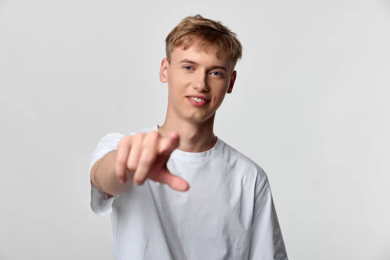 Pointing, man, gesture, casual, white tshirt, studio scene, direct outreach, engagement and invitation, confident expression, friendly contact for audience in marketing and social media campaigns