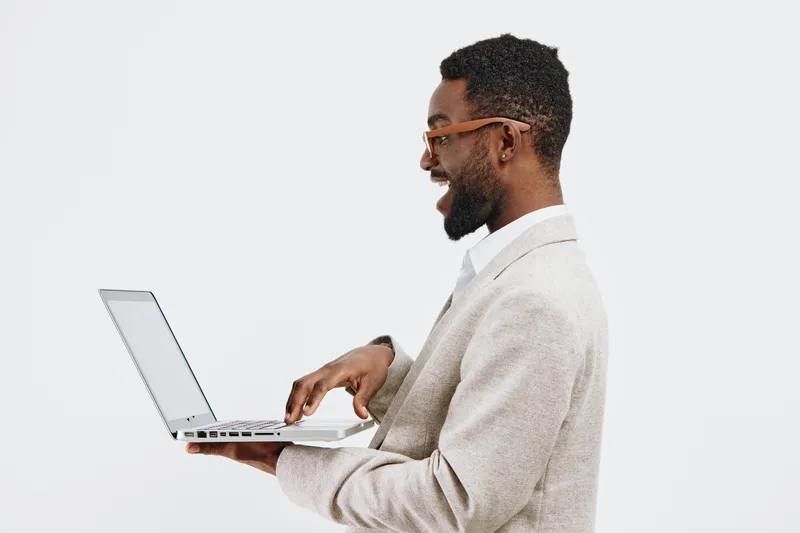 Smiling young African American man in business casual attire using laptop against plain white background. Studio shot, technology, communication, professional, desktop, modern lifestyle, confident.