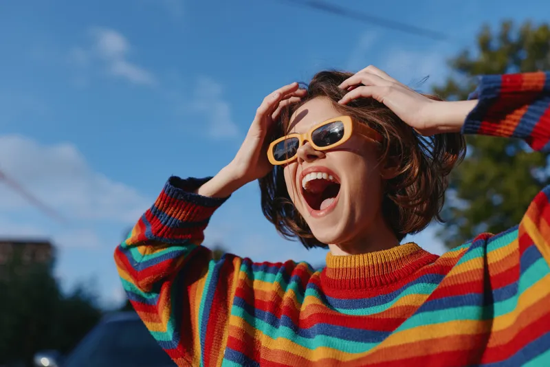 woman young woman striped sweater yellow sunglasses laughing outdoor joyful short hair casual outfit street fashion, young woman in colorful knit laughing with hands in hair for lifestyle travel