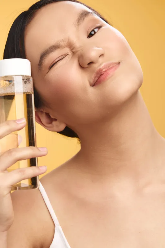 Asian woman is seen holding a water bottle in a bright studio setting. She smiles and tilts her head slightly, conveying refreshment, vitality, and a healthy lifestyle.