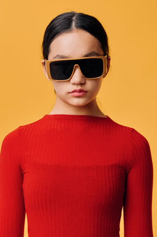 Woman wearing large sunglasses and red ribbed sweater posing against orange background with serious expression and sleek hair tied back in studio portrait.