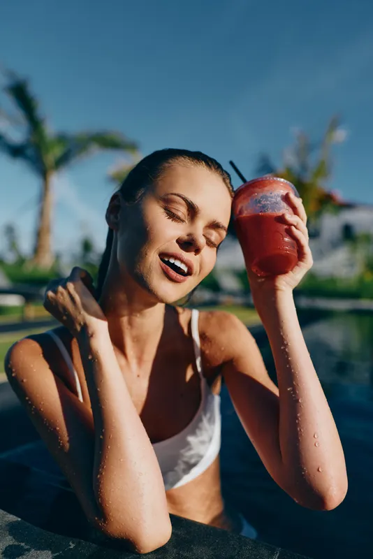 Young woman in white bikini relaxes in pool holding red smoothie cup with straw eyes closed enjoying sunny day tropical background lifestyle concept