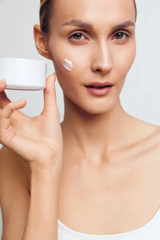 A young woman with clear skin applies moisturizing cream on her cheek while holding a white jar. The close-up shows natural beauty and skincare routine concept for healthy skin.