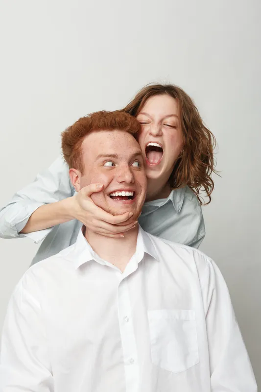 Playful young woman with curly hair hugs smiling redhead man from behind, both wearing white shirts on light background. Concept of friendship, fun, happiness, and joyful human connection.