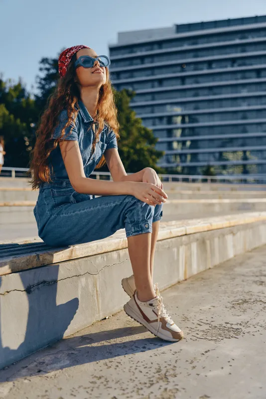 Lifestyle woman in modern boho-western denim street style wearing sunglasses and sneakers, relaxing outdoors in warm sunlight with film color tones enhancing mood.