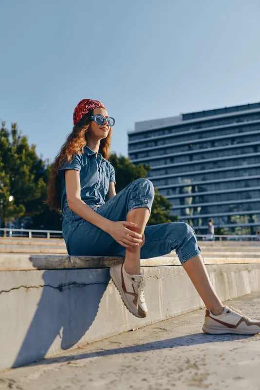 young woman sitting outdoors wearing denim jumpsuit and red headscarf with sunglasses relaxing against urban backdrop in sunny weather