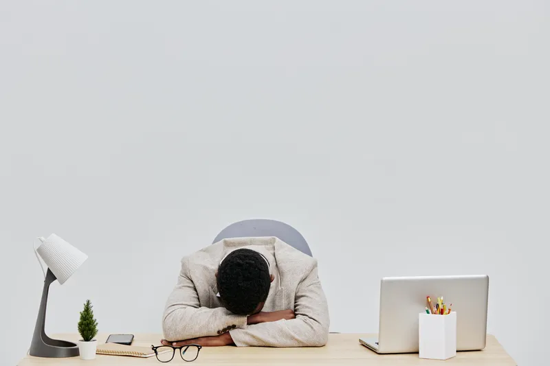 Man appears exhausted or frustrated, resting his head on folded arms on a clean, minimalist office desk with white background and modern workspace items, symbolizing work fatigue or stress. People lif