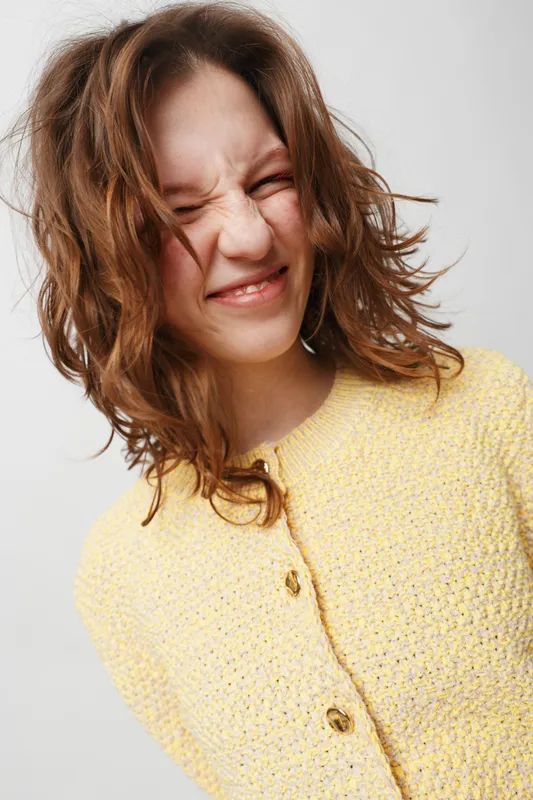 Young woman with messy hair wearing yellow knitted sweater makes a funny grimace with closed eyes on light background. Concept of playful mood, casual style, and natural expression.