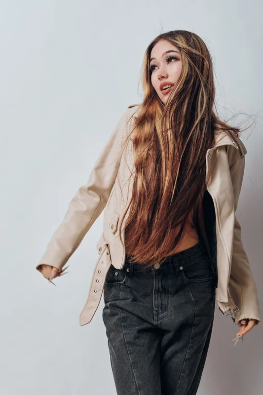 Young woman with long hair wearing beige jacket and black jeans posing on light background. Female model with casual fashion style standing isolated in studio with natural expression.