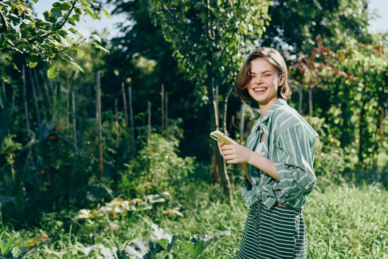 A cheerful woman stands in a lush garden, smiling while holding a device, wearing a striped shirt and skirt, bathed in warm sunlight.
