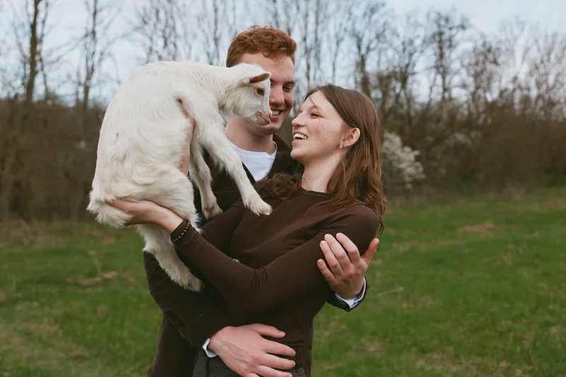 Cheerful couple outdoors holding a playful goat kid, smiling and embracing it, sunny moment in an open field, light breeze, natural colors, candid lifestyle scene with warm glow and carefree attitude
