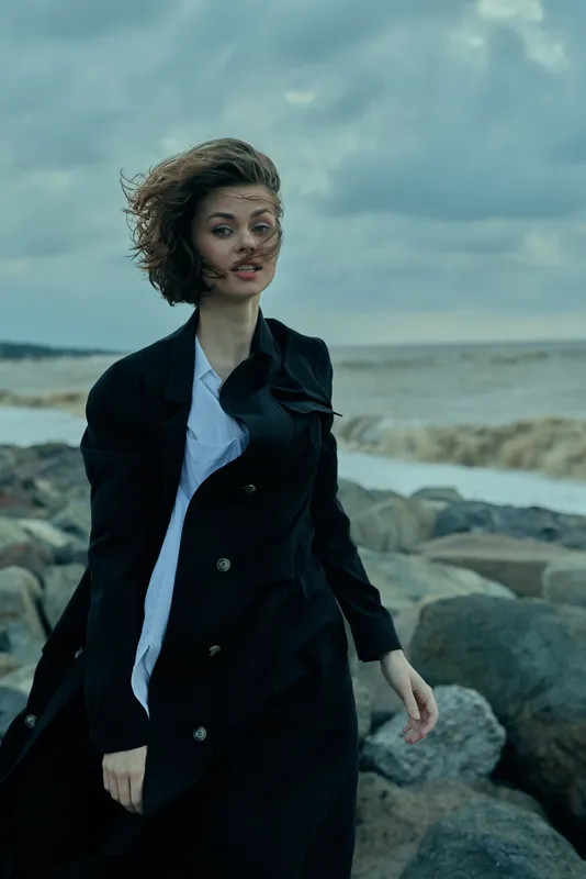 Windy coastal scene featuring a woman in a dark trench coat on a windy shore, waves crash, dramatic sky, motion and style in a moody outdoor moment with seascape grit
