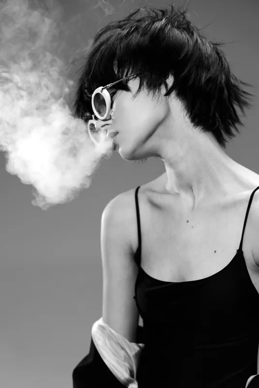 Black and white woman wearing sunglasses blowing smoke in stylish pose with short hair and black dress