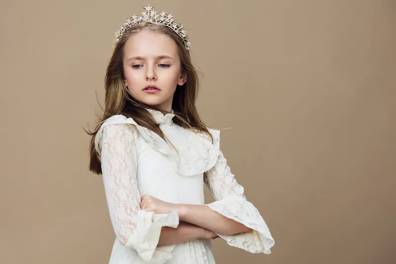 A poised young girl dressed in an elegant white dress with lace sleeves, wearing a crown, showcasing a confident expression against a soft beige background