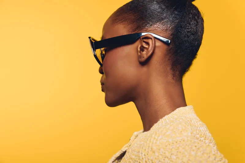Profile of a young black woman with dark skin wearing large black glasses and a yellow textured sweater against a bright yellow background. Concept of style and modern fashion.