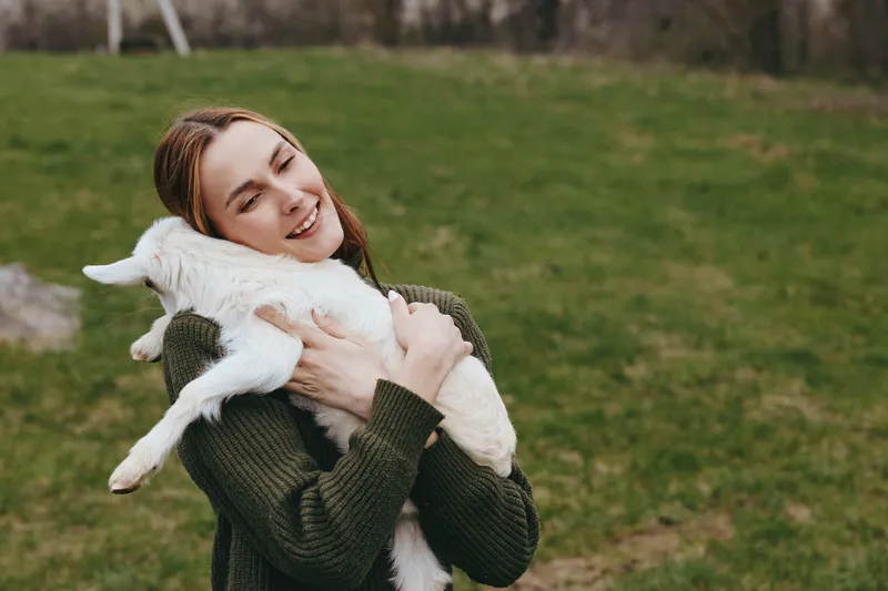 Joyful woman outdoors hugs a fluffy goat kid, sharing a tender moment in a grassy field, casual sweater, peaceful countryside mood, candid lifestyle scene, gentle interaction with animals.