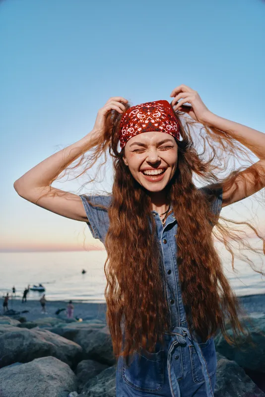 lifestyle woman with long hair wearing modern boho-western denim street style and red bandana, smiling joyfully outdoors at sunset with film color tones
