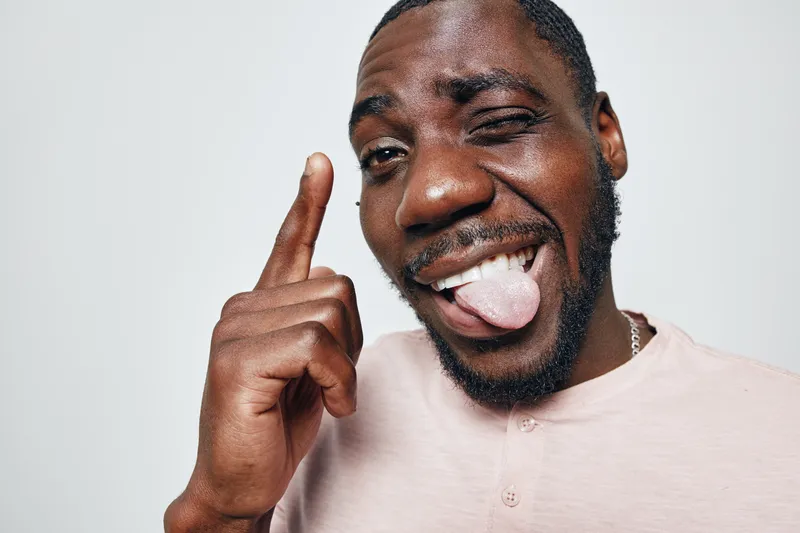 African American man with dark skin making funny face with tongue out and one eye closed, pointing finger up, casual shirt and beard on white background in studio.