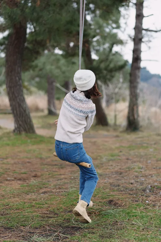 Person on a swing in a forest park, wearing a knit sweater, jeans and a beanie, enjoying a playful moment among tall trees and pine needles on the ground