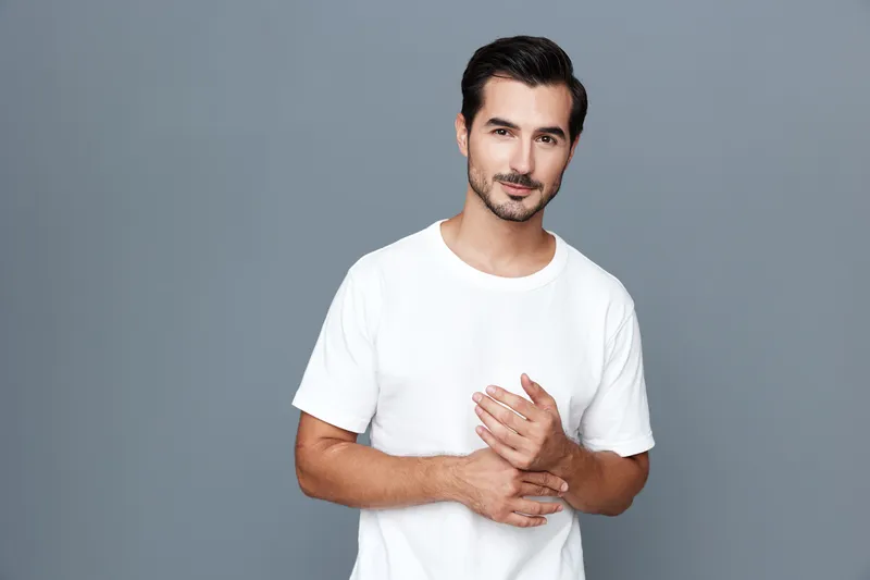 Smiling man standing confidently in a casual white t-shirt on a solid gray background, looking at the camera with a warm expression. People lifestyle concept.