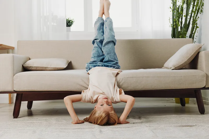Playful child in casual clothes doing headstand on carpet near sofa in bright living room. Concept of childhood fun, home activity, and energetic lifestyle.