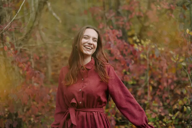 Candid portrait of a smiling young woman in autumn woods, genuine expression and natural pose conveying authentic mood and realistic warm tones, soft light, textured foliage and casual dress.