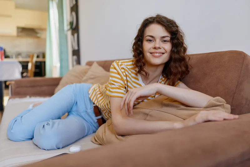 Casual woman relaxing at home on a sofa, smiling softly, enjoying quiet downtime in a cozy living room, dressed in striped top and jeans, natural light highlights comfort and warmth