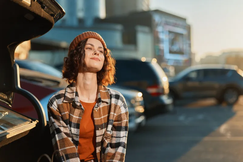woman portrait golden hour candid car parking beanie smile young adult sitting by open trunk, relaxed in plaid jacket, authenticity and candid lifestyle, golden hour glow, mindful living.
