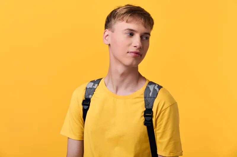 Student with backpack wearing a yellow shirt stands against a bright yellow backdrop, conveying curiosity and casual style suitable for education, youth, and everyday fashion contexts.