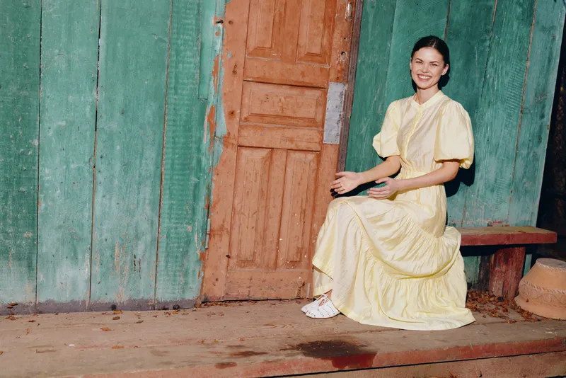 Woman in a pale yellow dress sits on a wooden bench beside a weathered doorway, against a turquoise wall, creating a calm rustic outdoor portrait.