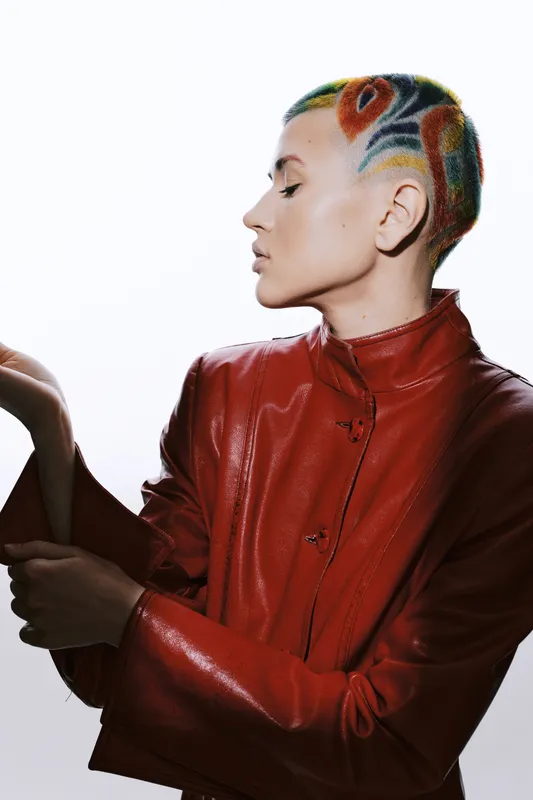 Woman with colorful short hair wearing a shiny red jacket poses confidently against white background, showcasing vibrant hairstyle and high-fashion urban look.