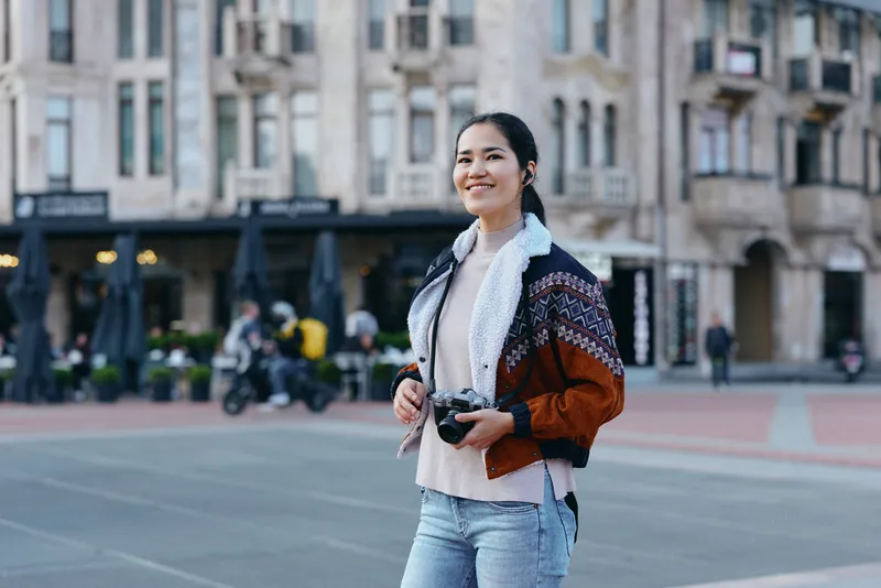 Woman with a camera on an urban street, wearing a cozy jacket and denim, smiling as she explores city life and captures candid moments and architecture.