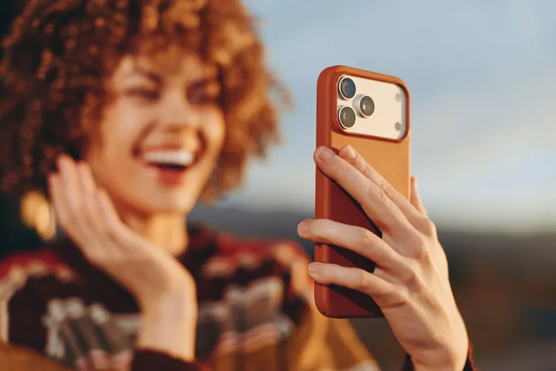 Woman holds smartphone for a selfie outdoors, lifestyle moment in summer sunlight, smiling and laughing, hand on phone camera, blurred urban background bokeh, casual modern portrait, youth