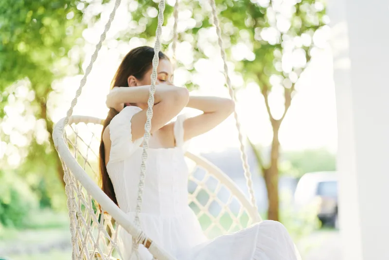 A serene young woman in a white dress sitting in a swing, enjoying a peaceful moment among green trees, conveying emotions of relaxation and tranquility