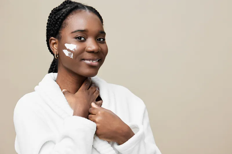 Beautiful Black woman applying skincare cream while wearing a cozy white robe, showing a radiant and confident smile in a neutral toned setting