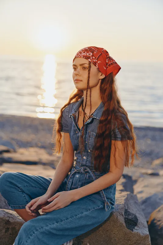 Lifestyle woman in modern boho-western denim street style with long hair and red bandana sitting by the sea during golden hour in warm film color tones.