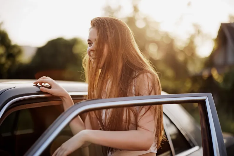A joyful young woman with long hair smiles while leaning out of a vintage car during sunset, capturing a carefree moment in nature's beauty.