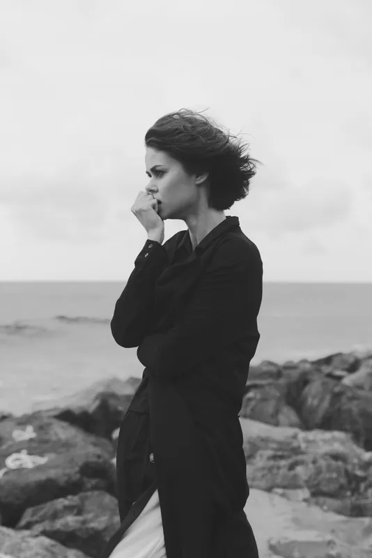 Woman in a dark coat standing on a rocky coastline by the sea, wind blown hair, thoughtful pose, moody monochrome scene ideal for dramatic lifestyle visuals