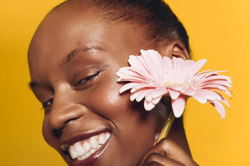 Close-up of happy African woman with dark skin smiling and holding pink flower near face on yellow background. Concept of natural beauty, skincare, wellness, and positive mood.