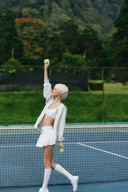 Energetic young woman in sporty attire with short blonde hair, playing tennis outdoors, vibrant green background, showcasing joy and athleticism