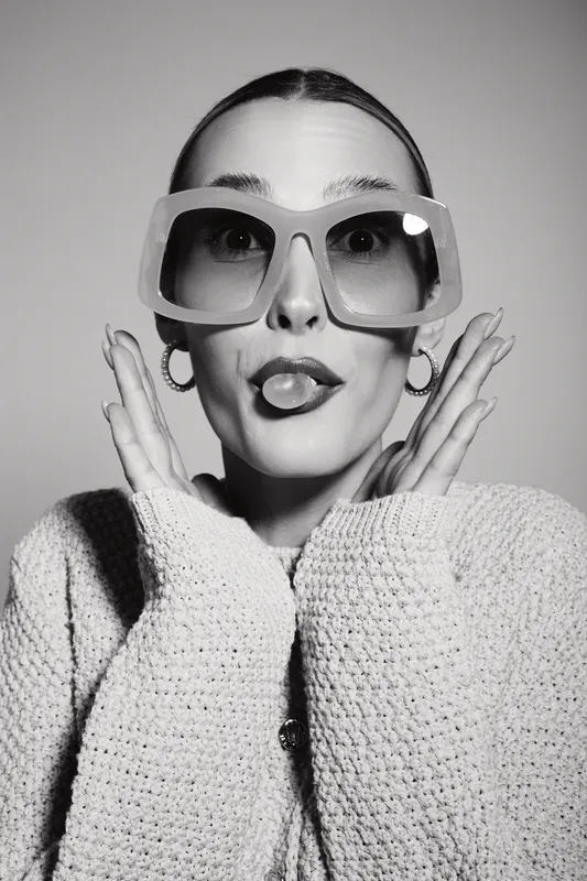 Young white woman in oversized sunglasses and knit sweater makes playful face with tongue out. Studio portrait in black and white. Concept of fun, fashion, and personality expression.