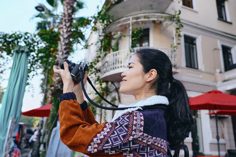 Photography of a woman photographer in a patterned sweater capturing urban street scenes with a camera, beside balconies and red umbrellas, showcasing travel, creativity, and street style.