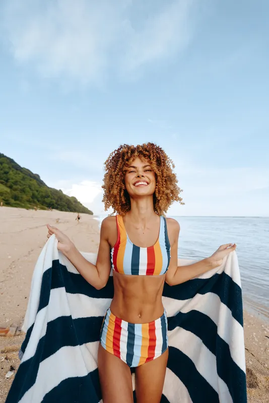 Happy young woman with curly hair in colorful striped bikini holds striped towel on sandy beach with ocean and green hill in background under clear blue sky.