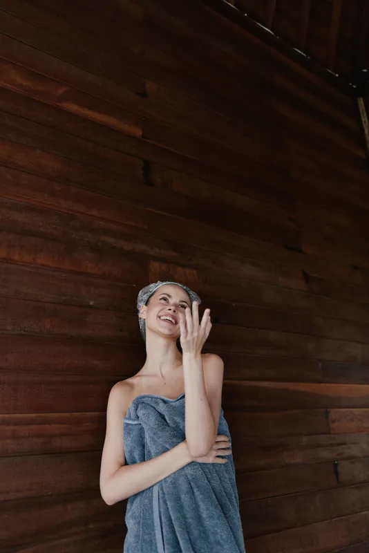 young woman wrapped in towel smiling joyfully after shower with towel turban, wooden wall background, natural light and relaxed mood