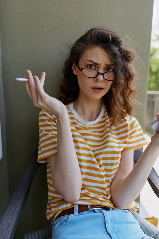 Young woman with striped shirt sits on a balcony, smoking a cigarette and glancing over her glasses with a curious, self amused expression under soft daylight.