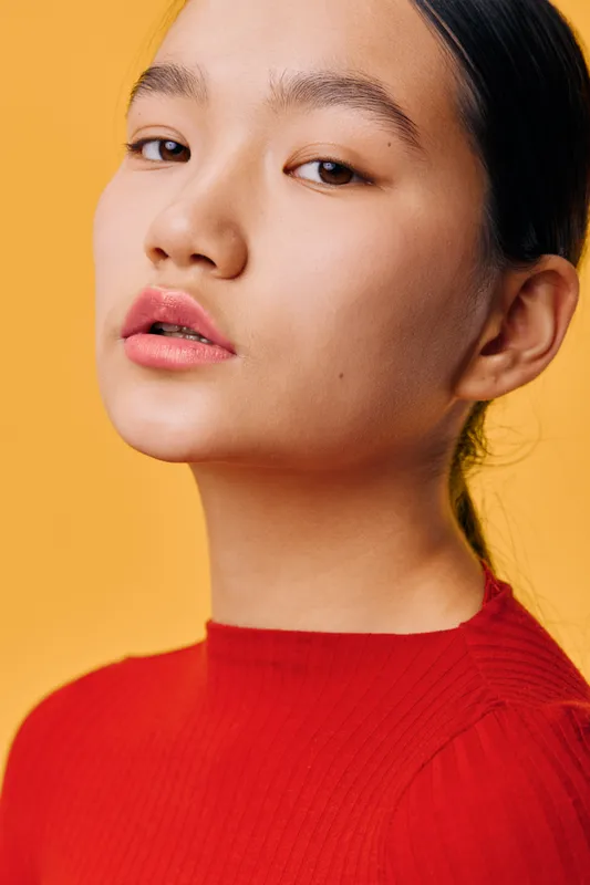 Portrait of young woman in red sweater with smooth skin and natural makeup, closeup on face and confident expression against orange background in studio with soft lighting.