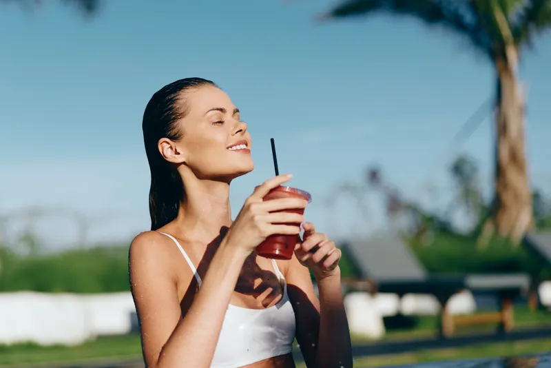 Bright outdoor portrait of a smiling woman in a white top, holding a red drink with a straw, enjoying sunny weather in a park with green grass and clear blue sky.