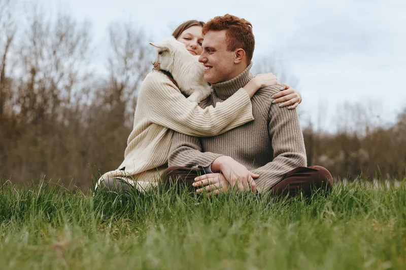 Couple shares a tender outdoor moment with a friendly goat kid, cuddling in cozy knitwear in a green field, bright sky overhead, calm laughter, and rustic charm highlighting affectionate connection