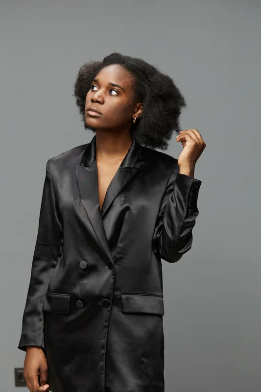 Confident young Black woman in a stylish black satin suit, looking upward with a thoughtful expression against a neutral gray background