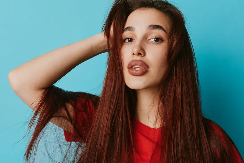 Woman with long red hair posing in red shirt against blue background looking at camera with hand in hair. Portrait of young female with natural makeup and confident expression in studio.